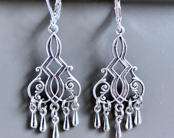 Peut inclure: Une paire de boucles d'oreilles argentées en forme de lustre. Chaque boucle d'oreille présente un motif orné avec un centre en forme de losange, des détails tourbillonnants et des breloques en forme de goutte. Fermoirs à levier.
