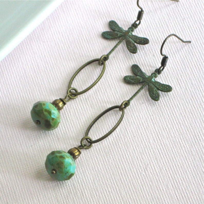 May include: A pair of dangle earrings featuring a turquoise green bead and a dragonfly charm. The earrings are made with brass and have a vintage look.