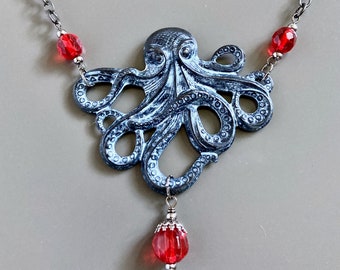 Op de afbeelding: Een zilveren ketting met een blauwe octopus hanger en rode glazen kralen. De octopus heeft een gedetailleerd ontwerp met textuur in de tentakels.