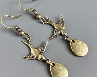 May include: A pair of antique bronze-colored earrings. Each earring features a detailed bird charm with a teardrop-shaped pendant. The earrings have a hook closure and a coiled spring detail near the top. The earrings are displayed against a gray background.