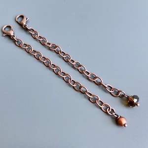 May include: Two copper chain extenders with lobster clasps. One extender has a brown crystal bead at the end, the other has a brown pearl bead.