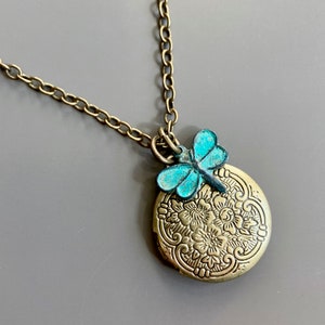 May include: A gold chain necklace with a round locket pendant featuring an intricate floral design. A turquoise dragonfly charm is attached to the top of the locket.