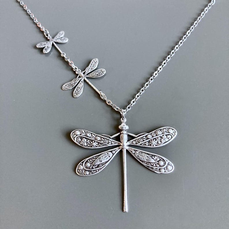 May include: Silver necklace with three dragonfly charms. The largest dragonfly charm is detailed with intricate floral patterns. The necklace is made of a delicate silver chain.