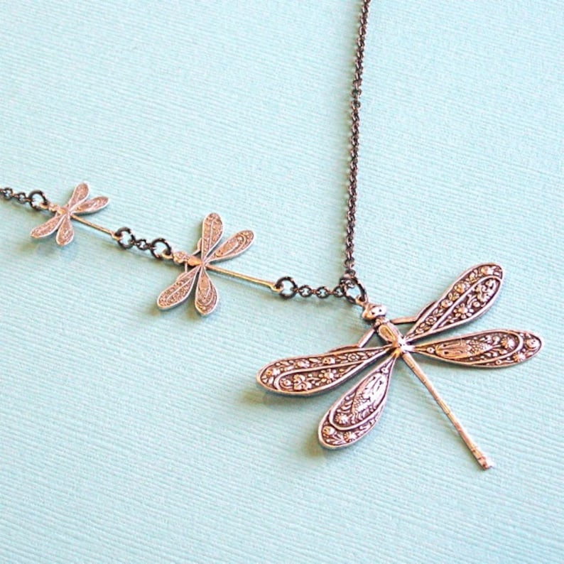 May include: A silver necklace with three dragonfly charms. The dragonflies are detailed with intricate patterns and have a vintage look.