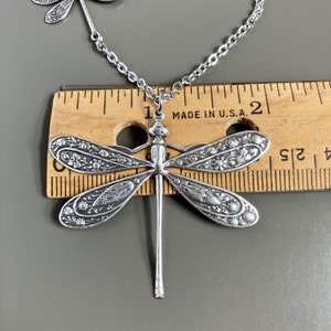 May include: Silver dragonfly pendant necklace with intricate floral details. The pendant is about 1 inch long and is hanging from a silver chain. The necklace is displayed on a wooden ruler with the text "MADE IN U.S.A."