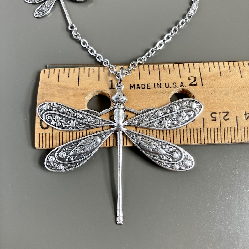 May include: Silver dragonfly pendant necklace with intricate floral details. The pendant is about 1 inch long and is hanging from a silver chain. The necklace is displayed on a wooden ruler with the text "MADE IN U.S.A."