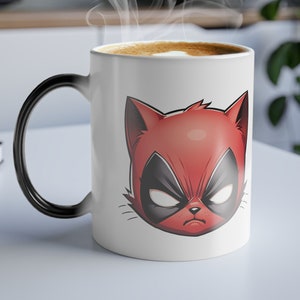 Puede incluir: Una taza de cerámica blanca que cambia de color con un diseño de cara de gato de dibujos animados rojo y negro. El asa de la taza es negra y el interior muestra un líquido marrón claro con vapor ascendente. La taza está sobre una superficie blanca.