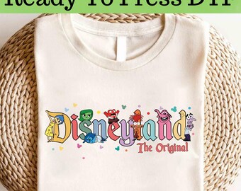 Op de afbeelding: Crèmekleurig T-shirt met de tekst "Ready To Press DTF" bovenaan. Het shirt toont het woord "Disneyland" in kleurrijke letters met cartoonfiguren en de woorden "The Original" eronder.