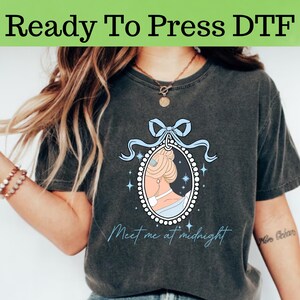 May include: A dark gray t-shirt with a graphic of a woman in a blue dress and a pearl necklace. The graphic is in a frame with a blue ribbon and the text "Meet me at midnight".