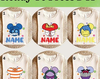 Op de afbeelding: Zes crèmekleurige t-shirts met cartoonkarakterontwerpen. Elk shirt heeft een ander karakter met een uniek kleurenschema en het woord "NAME" eronder. De tekst "Ready To Press DTF" staat bovenaan.