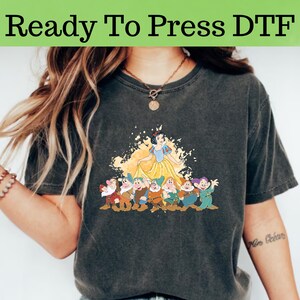 Op de afbeelding: Een donkergrijs T-shirt met een print van Sneeuwwitje en de Zeven Dwergen. De print heeft een gele en witte aquarel achtergrond met spatten verf. De tekst "Ready To Press DTF" staat bovenaan de afbeelding.