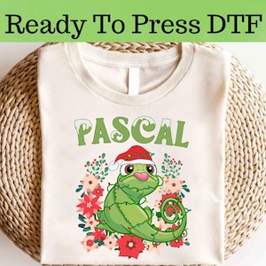 May include: A cream-coloured t-shirt with a green cartoon chameleon named Pascal wearing a Santa hat and wrapped in Christmas lights. The word "PASCAL" is printed above the chameleon in green letters. The shirt is surrounded by red and pink flowers.