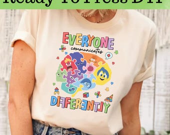 Op de afbeelding: Crèmekleurig T-shirt met de tekst "EVERYONE communicates DIFFERENTLY" in kleurrijke letters. Het ontwerp bevat een hersenvormige puzzel met verschillende cartoongezichten en andere kleine afbeeldingen.