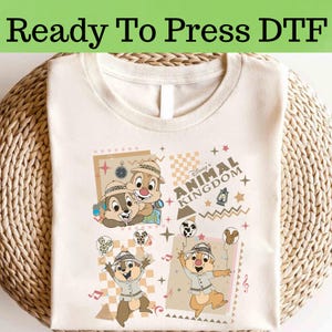 May include: Cream t-shirt with a colourful graphic of Chip and Dale from Disney's Animal Kingdom. The design includes the text "Disney's Animal Kingdom" and other decorative elements. The shirt is folded on a woven surface.