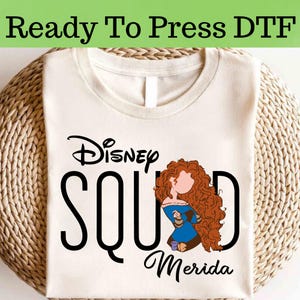 以下が含まれることがあります： 黒い文字で「Disney Squad Merida」と書かれたクリーム色のTシャツ。ディズニー映画「メリダとおそろしの森」のメリダの漫画イラストがデザインされています。「Ready To Press DTF」の文字が上部にあります。
