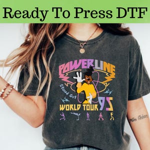 Puede incluir: Camiseta gris oscuro con un gráfico colorido de un personaje de dibujos animados y el texto "POWERLINE WORLD TOUR 95". La camiseta también tiene el texto "Ready To Press DTF" en la parte superior. El diseño incluye las palabras "Stand Out".