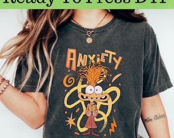 Op de afbeelding: Een donkergrijs T-shirt met een cartoonfiguur dat een rood-oranje jurk draagt. Het figuur heeft oranje haar en is omringd door gele swirls. De tekst "ANXIETY" is boven het figuur gedrukt.