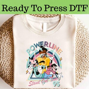 Puede incluir: Camiseta color crema con un gráfico colorido de personajes de dibujos animados, el texto "POWERLINE" arriba y "Stand Out Tour 95" abajo. La camiseta está lista para planchar.