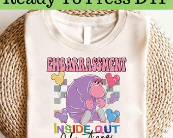 Op de afbeelding: Crèmekleurig T-shirt met een afbeelding van het personage Embarrassment uit de film Inside Out. Het ontwerp bevat het woord "EMBARRASSMENT" in roze, de tekst "INSIDE OUT" en de naam "Christiana". Het shirt is gelabeld "Ready To Press DTF".