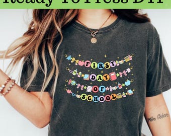 May include: Dark gray t-shirt with the text "Ready To Press DTF" at the top. The shirt features a colorful design that says "First Day Of School" with school-themed graphics like apples, books, and pencils.