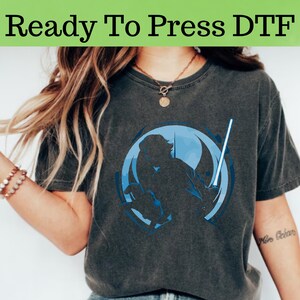 以下が含まれることがあります： 青いグラフィックが特徴の濃い灰色のTシャツ。ライトセーバーを持った人物が円形のデザインの中に描かれています。「Ready To Press DTF」というテキストが画像上部にあります。