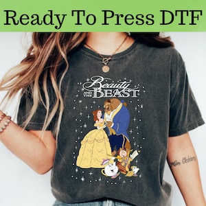 May include: A gray t-shirt with a vintage-style design featuring Belle and the Beast from Disney's Beauty and the Beast. The text "Beauty and the Beast" is written in white above the characters. The design also includes stars and other characters from the movie.