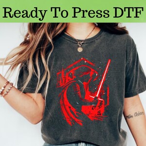 以下が含まれることがあります： 暗いグレーのTシャツに、ライトセーバーを持ったスターウォーズのキャラクターの赤いグラフィックがプリントされています。グラフィックは白いアウトラインと星で囲まれています。画像の上部に「Ready To Press DTF」というテキストがあります。