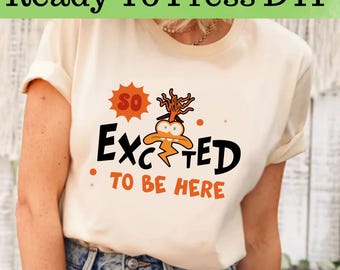 Op de afbeelding: Een wit T-shirt met een cartoonfiguur en de tekst "So Excited To Be Here".