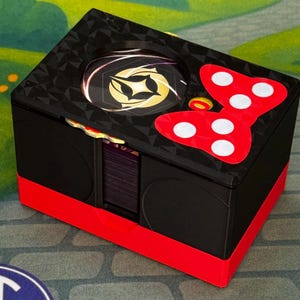 May include: A black box with a red base and a red bow with white polka dots on top. The box has a gold and black design on the top and a circular design in the center. The box is sitting on a gray surface with a blue and white logo.