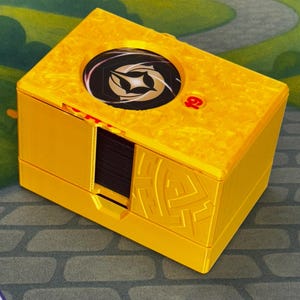 May include: A yellow card storage box with a black and white design on the top. The box has a textured surface and a sliding compartment. The background features a whimsical garden scene with a castle.