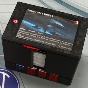 May include: Black card storage box with red and blue accents. The box has a card with the text "ORBITAL DOCK THETA-7" and a graphic of a spaceship. A Navycow Designs logo is visible on the mat.