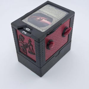 May include: A black and dark red rectangular deck box for trading cards featuring a textured finish. The top displays a Samwise the Stouthearted card under a clear cover. The side shows a black engraved silhouette and two red protruding accents.