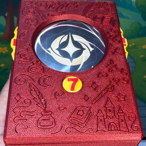May include: A red, rectangular box with a circular emblem and the number 7. The box has embossed designs of a quill, inkwell, book, wand, and castle. The emblem is a circular design with a star and geometric shapes.