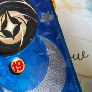 May include: Close-up of a blue, geometric-patterned object with a silver crescent moon and star design. A circular emblem with a gold and black design is visible, along with a yellow button with the number 19 in red.