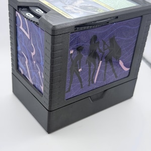 May include: A black, rectangular card storage box with a clear top. The sides feature a purple and pink design with black silhouettes of figures. The box has a sliding mechanism and a counter dial. A card is visible inside the box.