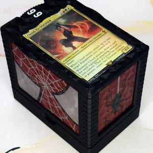 Puede incluir: Caja de almacenamiento de cartas negra con temática de Spider-Man. La caja presenta una carta con el texto "Cloud, Ex-SOLDIER" y el número "99". La caja tiene un diseño de telaraña roja y blanca en los laterales.