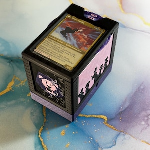 May include: A black and purple card storage box with a card featuring a spider-human hero. The box has a decorative design on the side and a lid. The card reads "Ghost-Spider" and includes game text. The box is designed to hold trading cards.