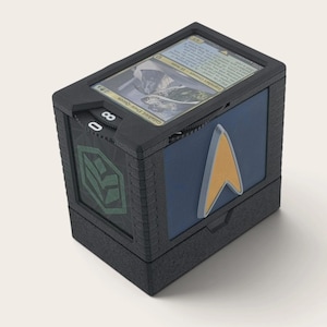 Peut inclure: Une boîte de jeu rectangulaire noire avec une fente sur le dessus pour une carte à collectionner, un cadran numérique latéral affichant le nombre 80 et un panneau latéral bleu orné d'un emblème doré en forme de delta. La boîte est texturée.