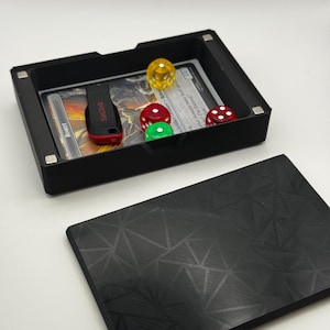 May include: A black rectangular game tray with a removable lid. Inside the tray are a USB drive, a game card, and several dice. The dice are red, green, and yellow. The lid has a geometric pattern.