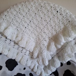Pode incluir: Um xale branco de crochê com uma borda com babados. O xale apresenta um delicado padrão de tecido aberto e é feito de um fio macio e texturizado. A borda com babados adiciona um toque decorativo ao acessório feito à mão.