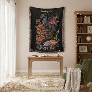 May include: A black woven throw blanket featuring a central illustration of a dragon perched on a stack of books surrounded by colorful flowers. The text "Evelyn's Reading blanket" is printed in gold script at the top and bottom of the design.