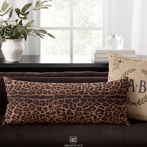 May include: A rectangular lumbar pillow featuring a brown and black leopard print pattern. The pillow is positioned horizontally on a dark brown sofa, showcasing a detailed animal-inspired design across its entire surface area.