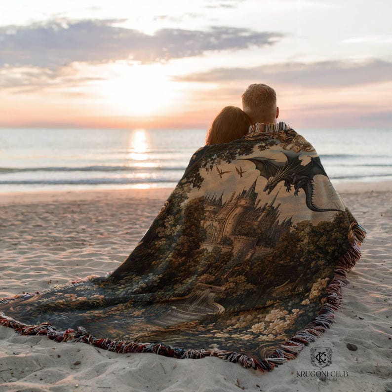 May include: A woven tapestry blanket featuring a castle, dragon, and forest scene. The blanket has a fringed edge and is draped over two people on a sandy beach at sunset. The blanket's design includes shades of brown, green, and gray.