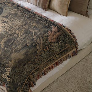 May include: A woven tapestry throw blanket featuring a detailed dark-toned landscape scene with architectural structures, lush foliage, and floral elements. The blanket has a decorative fringe border with multi-colored threads along the edges.