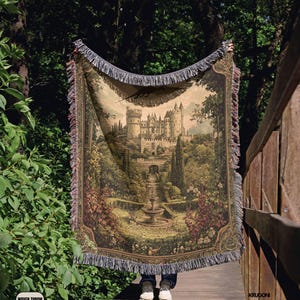 Pode incluir: Uma manta tecida com uma ilustra&ccedil;&atilde;o detalhada em estilo de tape&ccedil;aria de um castelo de pedra rodeado por jardins exuberantes, &aacute;rvores e uma fonte central. O tecido retangular tem bordas com franjas decorativas na parte superior e inferior.