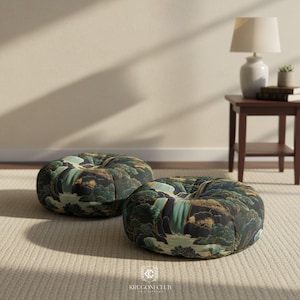 Two round floor pillows featuring a Japanese Hokusai-style waterfall print in deep blue, green, and earthy tones. The pillows are placed on a neutral beige carpet in a softly lit minimalist room, beside a wooden side table with a lamp and books.