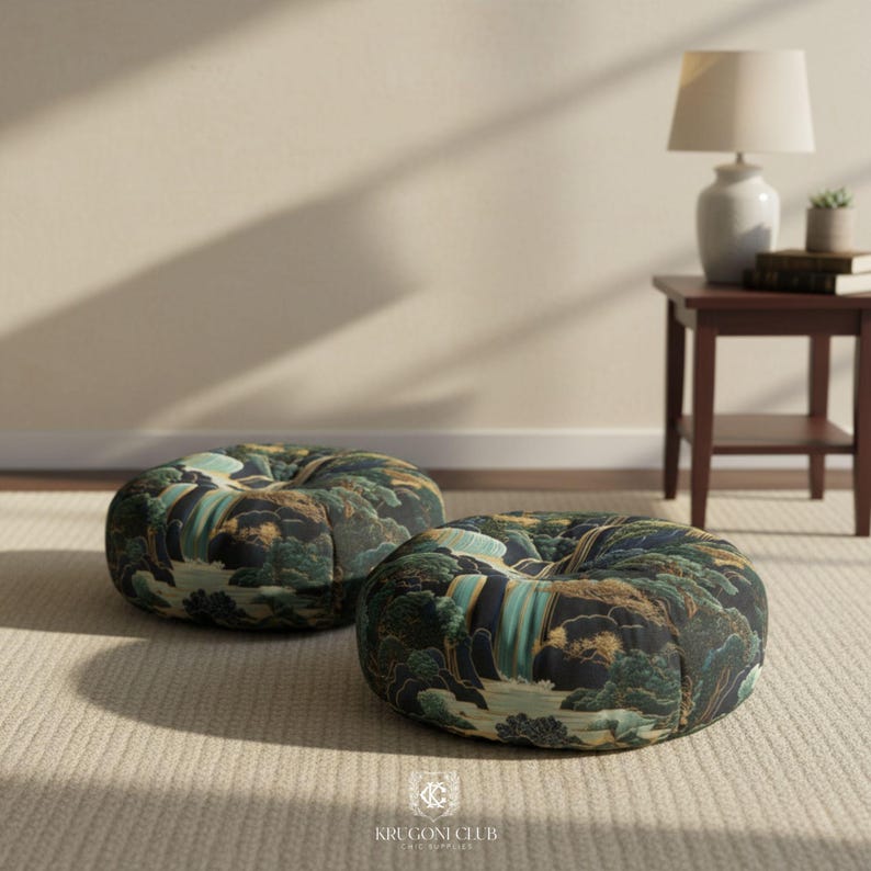 Two round floor pillows featuring a Japanese Hokusai-style waterfall print in deep blue, green, and earthy tones. The pillows are placed on a neutral beige carpet in a softly lit minimalist room, beside a wooden side table with a lamp and books.