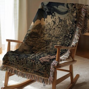 May include: A woven throw blanket featuring a detailed tapestry-style illustration of a large dragon flying over a stone castle and lush landscape. The blanket has a fringed edge and is draped over a wooden rocking chair with a light-colored seat.
