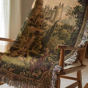 Pode incluir: Uma manta tecida com uma cena detalhada em estilo de tape&ccedil;aria de um castelo de pedra, um jardim exuberante com uma fonte e um drag&atilde;o voador. A manta tem uma borda decorativa e franjas, disposta sobre uma cadeira de balan&ccedil;o de madeira.