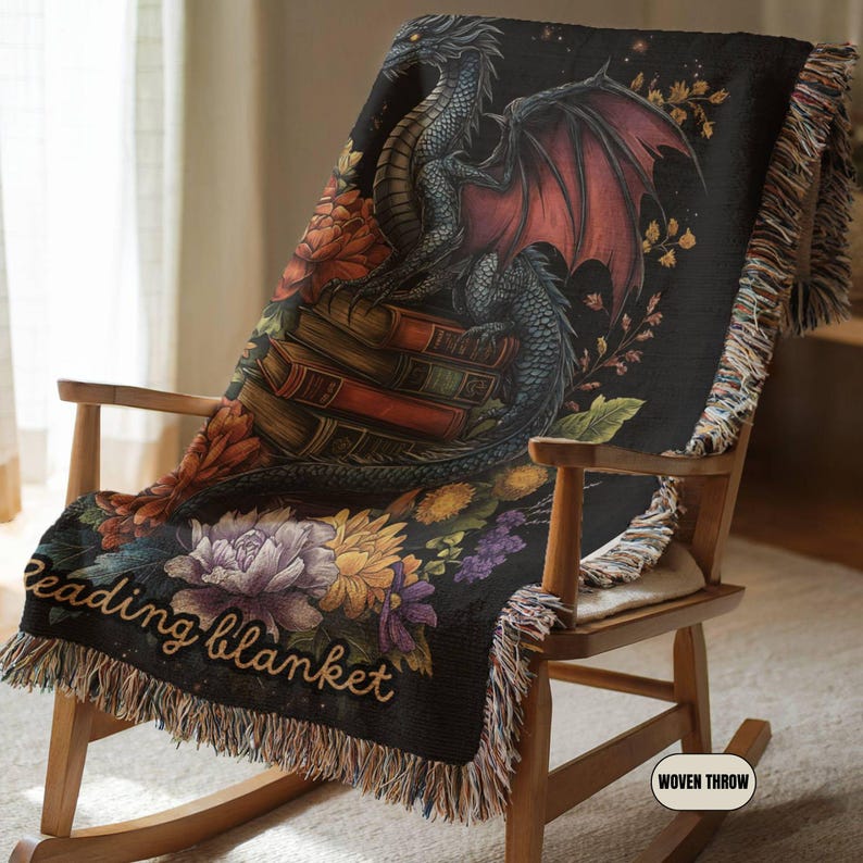 May include: A black woven throw blanket featuring a dragon perched on a stack of books, surrounded by colorful floral illustrations. The text "reading blanket" is printed in a script font at the bottom, and the edges have a decorative fringe.